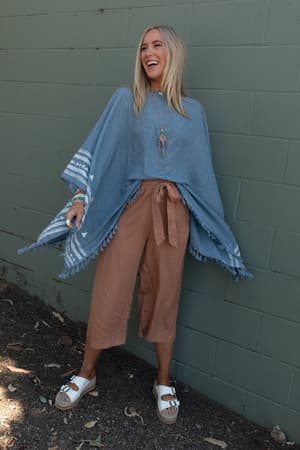 A smiling woman stands outdoors against a green brick wall, wearing the Fallon Embroidered Sleeve Tassel Poncho in blue with her arms spread wide.