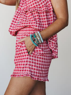 A close-up side shot of a red gingham and heart-print shorts set styled with a stack of turquoise and gold beaded bracelets — equal parts playful and sweet.
