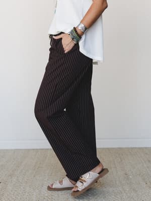 A side-profile shot of wide-leg pinstripe pants in deep chocolate brown, styled with a white cropped top, ivory buckle sandals, and a stacked arm of beaded and silver bracelets — relaxed and put-together at the same time.