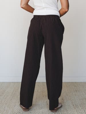 Shot from the back, a model wears wide-leg pinstripe trousers in a deep chocolate brown paired with a simple white ribbed top — relaxed and pulled-together in equal measure.
