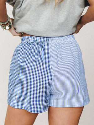 A mixed-print shorts moment that earns a second look — gingham on one side, seersucker stripes on the other, styled casually with a gray tee and stacked turquoise bracelets.