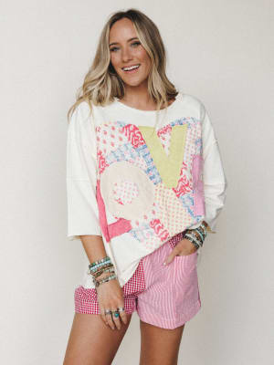 An oversized cream sweatshirt with a colorful patchwork 'LOVE' appliqué is paired with mixed-pattern red and white shorts — gingham and stripe panels — and stacked boho jewelry for a playful, lived-in spring look.