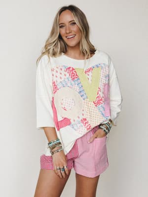 An oversized cream sweatshirt with a colorful patchwork 'LOVE' appliqué is paired with mixed-pattern red and white shorts — gingham and stripe panels — and stacked boho jewelry for a playful, lived-in spring look.