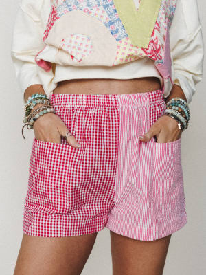 A playful close-up of mixed-print shorts in red gingham and pink stripe panels, styled with a patchwork sweatshirt and a full stack of beaded bracelets — the kind of outfit that has a very specific 'I dressed myself and I loved every second of it' en