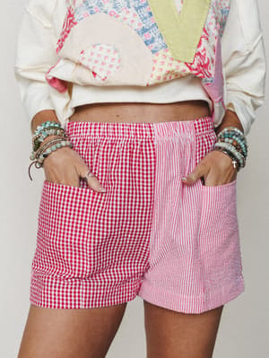 A playful close-up of mixed-print shorts in red gingham and pink stripe panels, styled with a patchwork sweatshirt and a full stack of beaded bracelets — the kind of outfit that has a very specific 'I dressed myself and I loved every second of it' en