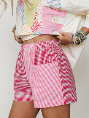 A close-up mid-shot of mixed red gingham and seersucker stripe shorts styled with a patchwork cream top and stacked beaded bracelets — playful, summery, and full of personality.