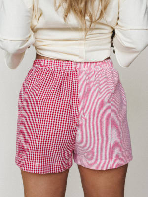 A back-view shot of mixed-print shorts pairing red gingham on one side with pink and white vertical stripes on the other — playful, a little unexpected, and styled casually with a knotted cream top.