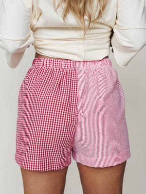 A back-view shot of mixed-print shorts pairing red gingham on one side with pink and white vertical stripes on the other — playful, a little unexpected, and styled casually with a knotted cream top.