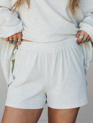 A close-up shot of a matching ribbed stripe set — the shorts are the star here, with a relaxed elastic waist and soft baby blue colorway, styled with stacked silver and turquoise rings that feel totally at home with the laid-back vibe.