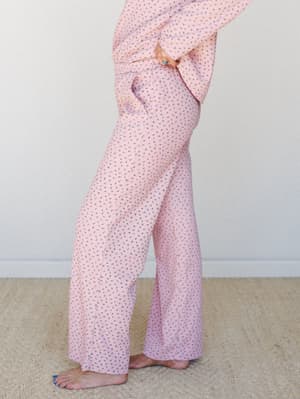 A side-profile mid-shot of wide-leg floral pants in soft pink with a matching top, styled barefoot against a neutral studio wall — cozy and a little dreamy, like a lazy Sunday that somehow looks pulled-together.