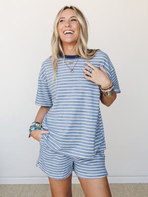 A laughing, sun-kissed model wears a relaxed striped tee-and-shorts matching set in soft blue and cream, styled with stacked boho bracelets and layered necklaces — the kind of outfit that makes a Saturday feel like a small vacation.