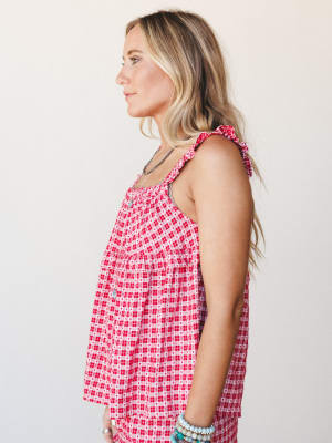 A side-profile shot of a blonde model wearing a flowy red gingham top with heart print detailing and ruffle tie straps, styled with layered silver and turquoise jewelry against a soft studio backdrop — sweet and summery with just enough personality.