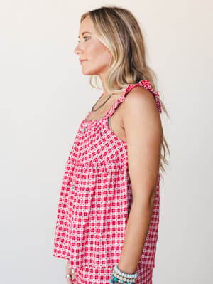 A side-profile shot of a blonde model wearing a flowy red gingham top with heart print detailing and ruffle tie straps, styled with layered silver and turquoise jewelry against a soft studio backdrop — sweet and summery with just enough personality.