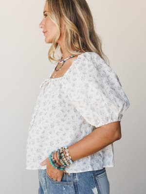 A breezy floral blouse in cream and dusty blue styled with denim and stacked beaded bracelets — the kind of top that feels like a good mood.