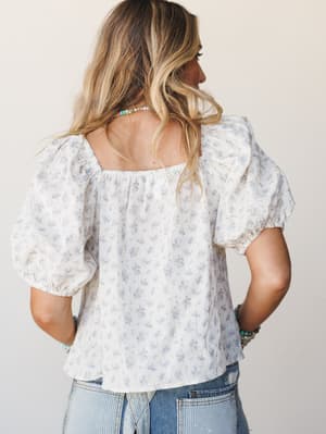 Shot from behind, a breezy floral blouse with voluminous puff sleeves and a square back neckline is paired with light wash jeans — the kind of top that makes a simple outfit feel like it had a little thought behind it.
