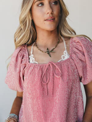 A close-up model shot of a red gingham puff sleeve top with lace trim neckline, styled with a brass bird pendant necklace and beaded bracelets — sweet, a little nostalgic, and very much a summer Saturday.