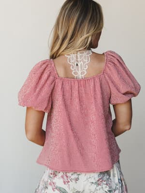 Shot from behind, a model wears a red gingham puff-sleeve top with a delicate lace back cutout detail — sweet, a little romantic, and styled with a floral skirt peeking in at the hem.