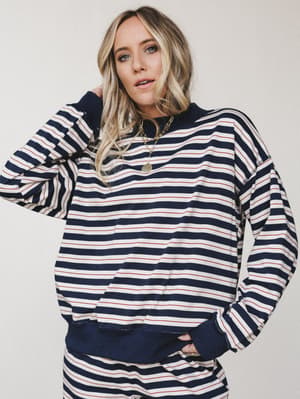 A relaxed striped pullover sweatshirt in classic navy, cream, and red styling — worn with layered gold necklaces and an effortless hand-in-pocket ease that feels more weekend coffee run than couch day.