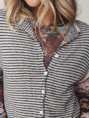 A close-up torso shot of a striped knit sweater vest layered over a floral print top, styled with gold chain necklaces — the kind of effortless pattern mixing that looks intentional without trying too hard.