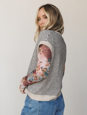 A model shot over-the-shoulder showing a striped sweater vest layered over a floral long-sleeve top — the kind of mix-and-match combo that looks like you meant to do it, and you did.