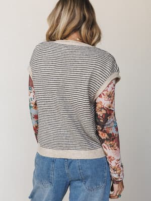 A back-view shot of a mixed-media top pairing a striped knit sweater vest over a floral long-sleeve underlayer, worn with medium-wash jeans — the kind of effortless layered look that feels intentional without trying too hard.