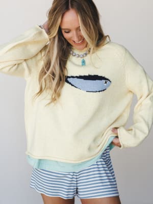 A smiling model in a butter yellow oversized knit sweater with a surfboard intarsia graphic layers over a mint top, paired with blue and white striped shorts — casual, coastal, and the kind of outfit that makes a Tuesday feel like vacation.