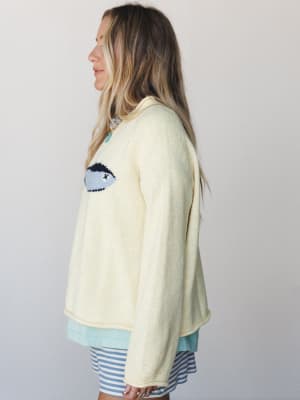 A cream knit sweater with a playful intarsia eye graphic is layered over a mint top and blue-and-white striped shorts, shot in profile against a soft studio wall — casual and a little artsy in the best way.