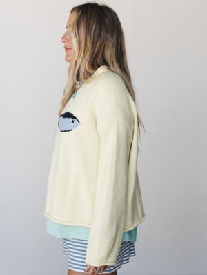 A cream knit sweater with a playful intarsia eye graphic is layered over a mint top and blue-and-white striped shorts, shot in profile against a soft studio wall — casual and a little artsy in the best way.