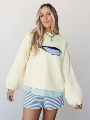 A cream knit pullover with a surfboard intarsia graphic is layered over a mint top and paired with blue striped shorts — casual, coastal, and the kind of outfit you'd actually wear on a beach town coffee run.
