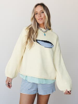 A cream knit pullover with a surfboard intarsia graphic is layered over a mint top and paired with blue striped shorts — casual, coastal, and the kind of outfit you'd actually wear on a beach town coffee run.