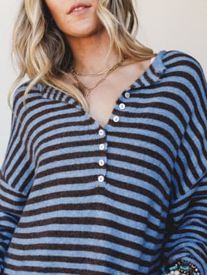 A cozy striped henley sweater top shot up close on a model, the blue and brown tones feeling effortlessly lived-in — layered with gold chain necklaces and a stack of beaded bracelets peeking in at the wrist.