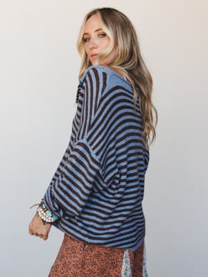A model glances over her shoulder in a relaxed striped linen top layered over a floral skirt, stacked with turquoise and pearl bracelets — the kind of effortless mix-and-match that looks accidental but totally isn't.