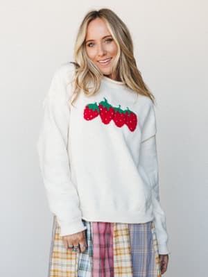 A cream knit pullover sweater with a playful intarsia strawberry graphic across the chest, styled with a multicolor plaid midi skirt — the kind of outfit that makes a Tuesday feel like a little event.