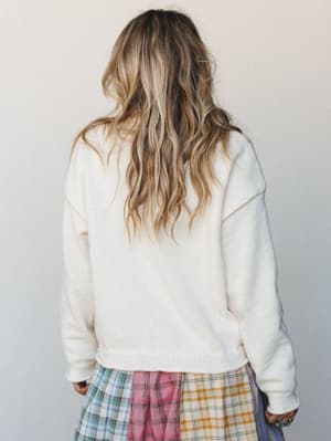 Shot from behind, a cream knit sweater pairs with a colorful mixed plaid skirt — soft and a little unexpected, the kind of outfit that looks like it came together effortlessly on a good-hair day.