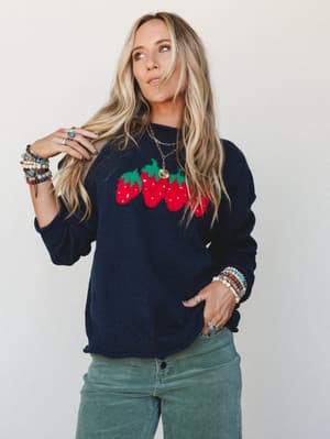 A navy knit sweater with a bold strawberry intarsia graphic worn over sage green corduroy wide-legs, styled with stacked boho bracelets and a gold chain necklace — the kind of outfit that feels intentional without trying too hard.