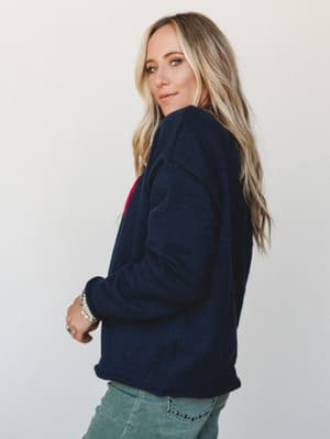 Shot from a three-quarter back angle, she glances over her shoulder in a cozy navy knit sweater with a glimpse of red graphic detail at the front — paired with sage green jeans and layered boho jewelry for an effortlessly lived-in, cool-girl vibe.
