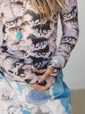 A close-cropped torso shot of a mesh long-sleeve top covered in a watercolor wild horse print, styled with graphic bleach-wash jeans and a turquoise pendant necklace — the kind of outfit that has a whole personality.