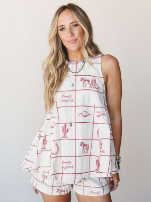A cream sleeveless swing top and matching shorts set printed with a red grid pattern and western embroidery motifs — horses, cowboy boots, cacti, and 'Howdy Cowgirls' script — styled with layered necklaces and turquoise bracelets for a playful, cowgi