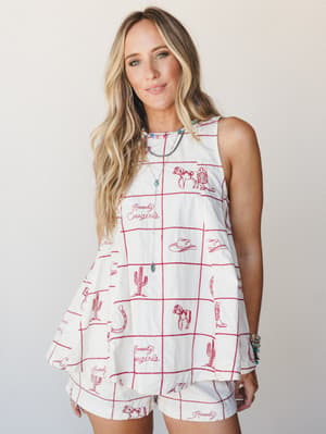 A cream sleeveless swing top and matching shorts set printed with a red grid pattern and western embroidery motifs — horses, cowboy boots, cacti, and 'Howdy Cowgirls' script — styled with layered necklaces and turquoise bracelets for a playful, cowgi