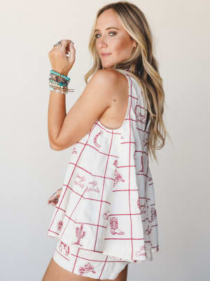 A breezy cream tank top with cherry red grid lines and western-themed novelty embroidery — cowboy boots, cacti, horseshoes, script text — styled with a stack of turquoise and beaded bracelets for an effortless western-boho feel.