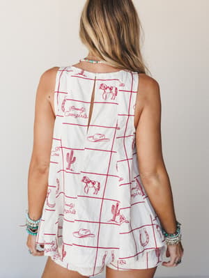 A breezy sleeveless top in cream with allover red 'Howdy Cowgirls' western novelty embroidery — horses, cacti, cowboy hats, horseshoes — shot from the back to show a keyhole detail, styled with layered turquoise and beaded jewelry.