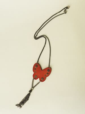 A rust-red butterfly pendant necklace with circular cutouts and a dangling chain tassel, laid flat against a warm cream background — the kind of piece that does all the talking.