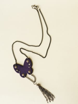 A long chain necklace laid flat on a cream surface, featuring a cutout purple butterfly pendant with circular perforations and a dangling gunmetal chain tassel — the kind of piece that does the talking for a simple outfit.