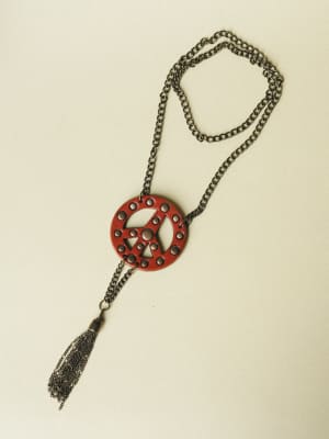 A long chain necklace laid flat on a neutral background, featuring a red leather peace sign pendant with circular cutouts and a dangling chain tassel — the kind of piece that makes a simple tee feel intentional.