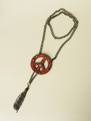 A long chain necklace laid flat on a neutral background, featuring a red leather peace sign pendant with circular cutouts and a dangling chain tassel — the kind of piece that makes a simple tee feel intentional.