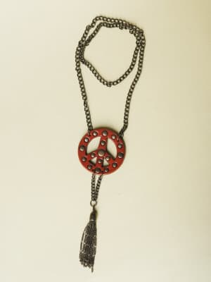 A long gunmetal chain necklace laid flat, centered on a cream background — the deep red leather peace sign pendant is studded with small cutout circles and finished with a dangling chain tassel that gives it an easy, free-spirited edge.