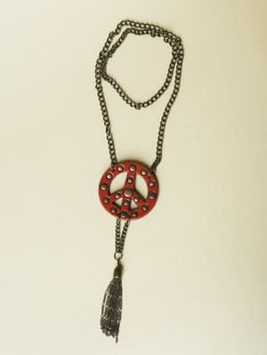 A long gunmetal chain necklace laid flat, centered on a cream background — the deep red leather peace sign pendant is studded with small cutout circles and finished with a dangling chain tassel that gives it an easy, free-spirited edge.