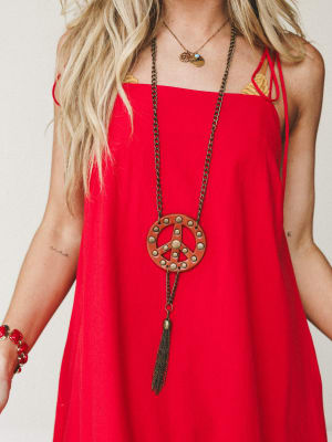 A bold red slip dress with tie shoulders is styled with a standout long bronze chain necklace featuring a studded leather peace sign pendant and tassel drop — the kind of look that has a whole personality before you even say hello.
