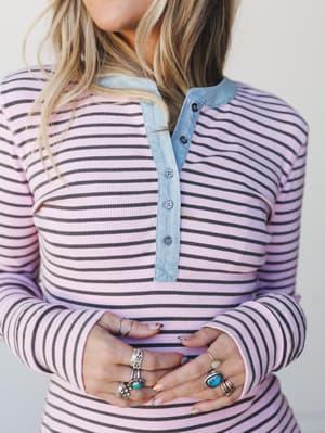 A close-up torso shot of a ribbed striped henley top in blush pink and dark brown, styled with layered silver and turquoise rings — the kind of top that looks like it's been in someone's rotation for years in the best way.