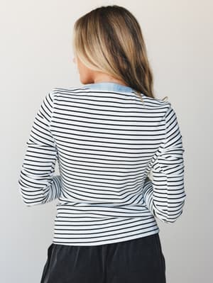 A back-view shot of a fitted ribbed stripe long-sleeve top layered over a light chambray shirt, paired with dark charcoal bottoms — easy, put-together, the kind of outfit that looks intentional without trying.